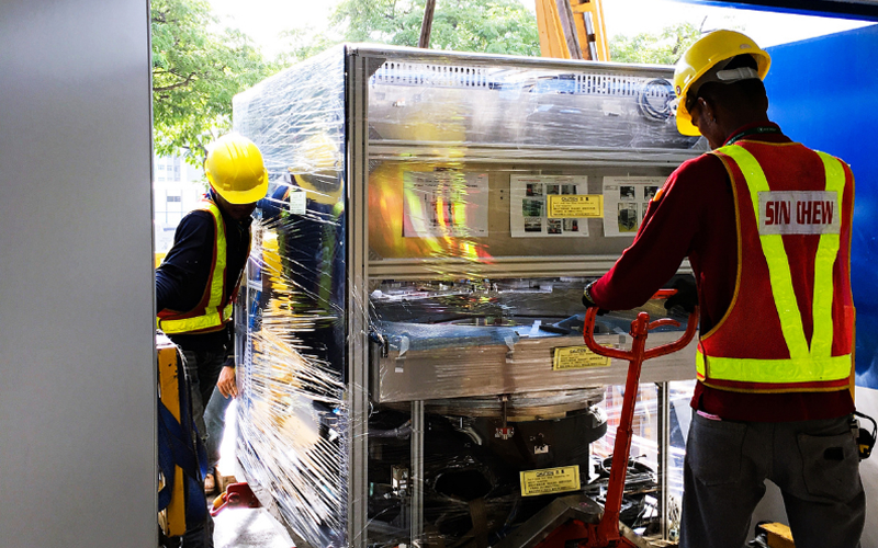 Logistics workers moving heavy machinery wrapped in protective plastic.